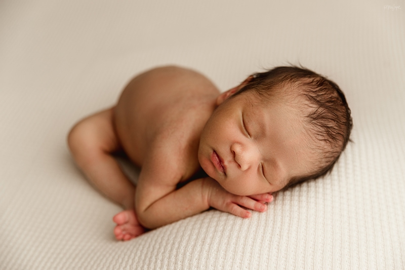 Newborn Photographer, a little baby lays on his arms while comfy on bed linens