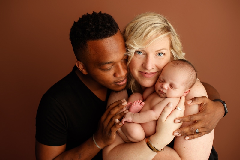 Newborn Photographer, mom holds baby as dad hugs mom and admires them both