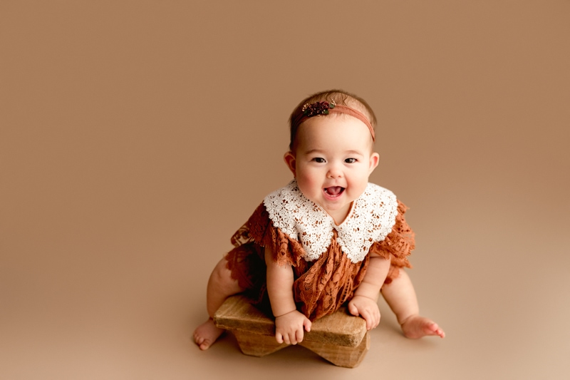 Baby Photographer, a little girl leans on a wooden stool smiling, she wears a dress