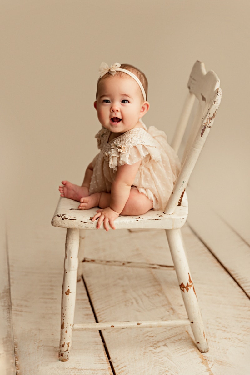 Baby Photographer, a baby girl sits on a wooden chair happy, she wears a dress and headband