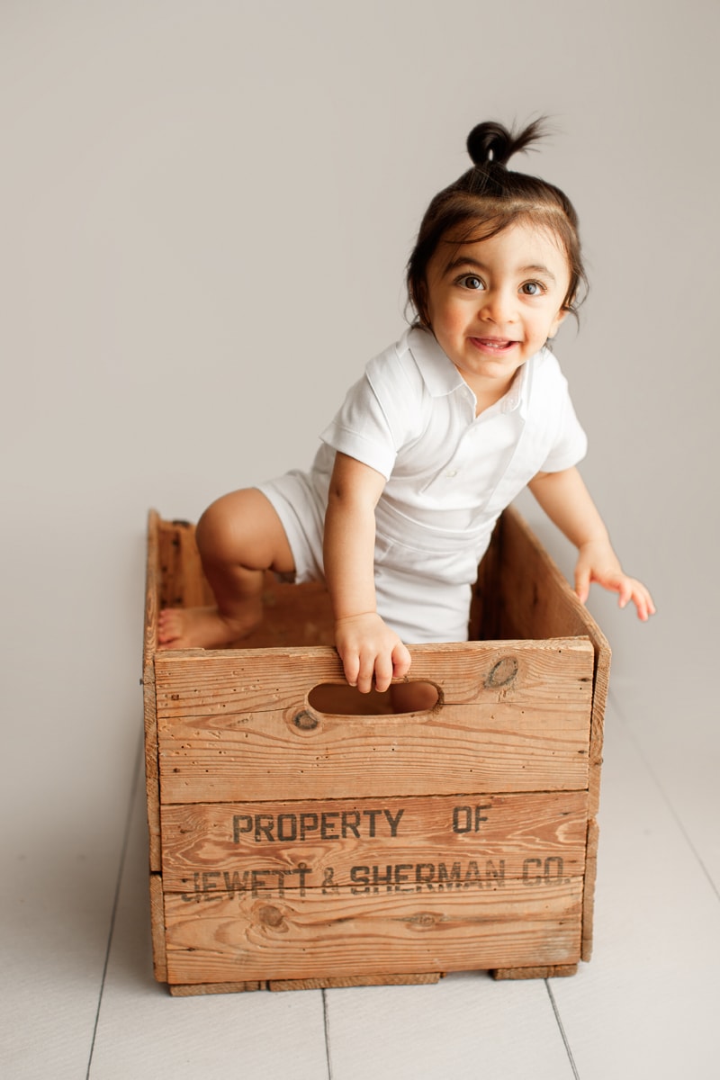 Baby Photographer, a young child climbs out of a wooden crate smiling