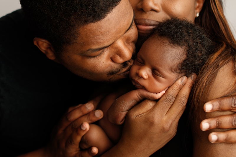 Newborn Photographer, a father kisses his baby son and squeezes mom tight at the same time