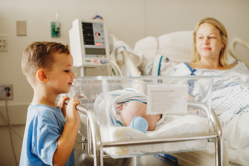 Fresh 48 Photographer, in a delivery room, older brother looks at his newborn sibling, mom looks on from delivery bed
