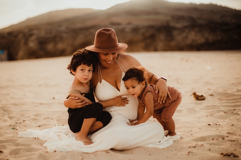 Family Photographer, a mother sits in the sand, her two young boys are happily huddling close to her