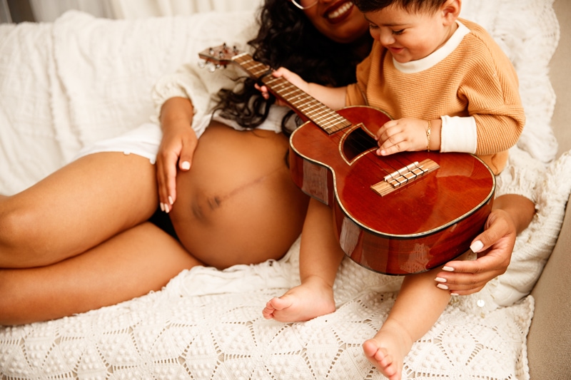 Fine Art Maternity Photographer, a mother places hand on belly as admires her son playing with a guitar beside her