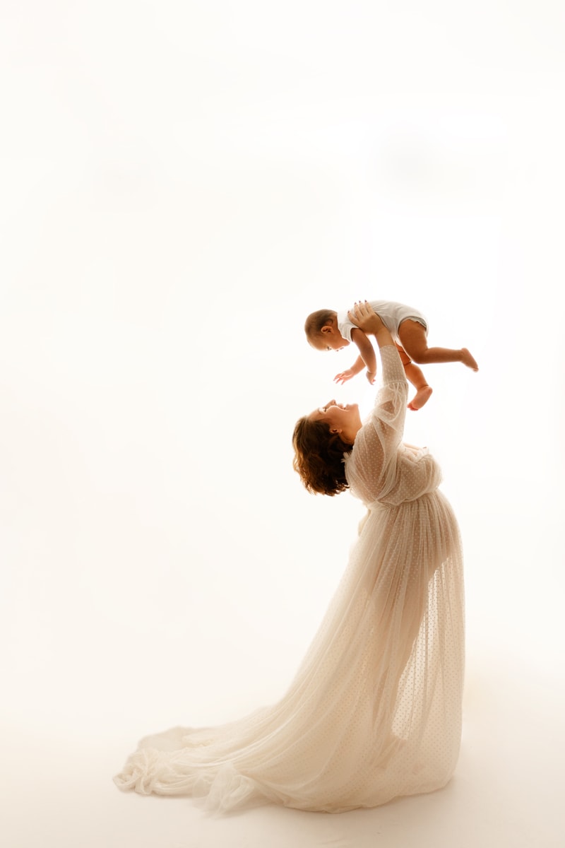 Fine Art Maternity Photographer, a pregnant mother wears a tule dress and swings her other baby in the air with smiles