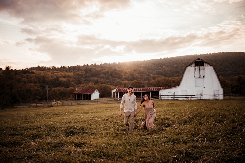 Family Photographer, a man and woman walk hand-in-hand through a grassy field, a farm stands tall behind them