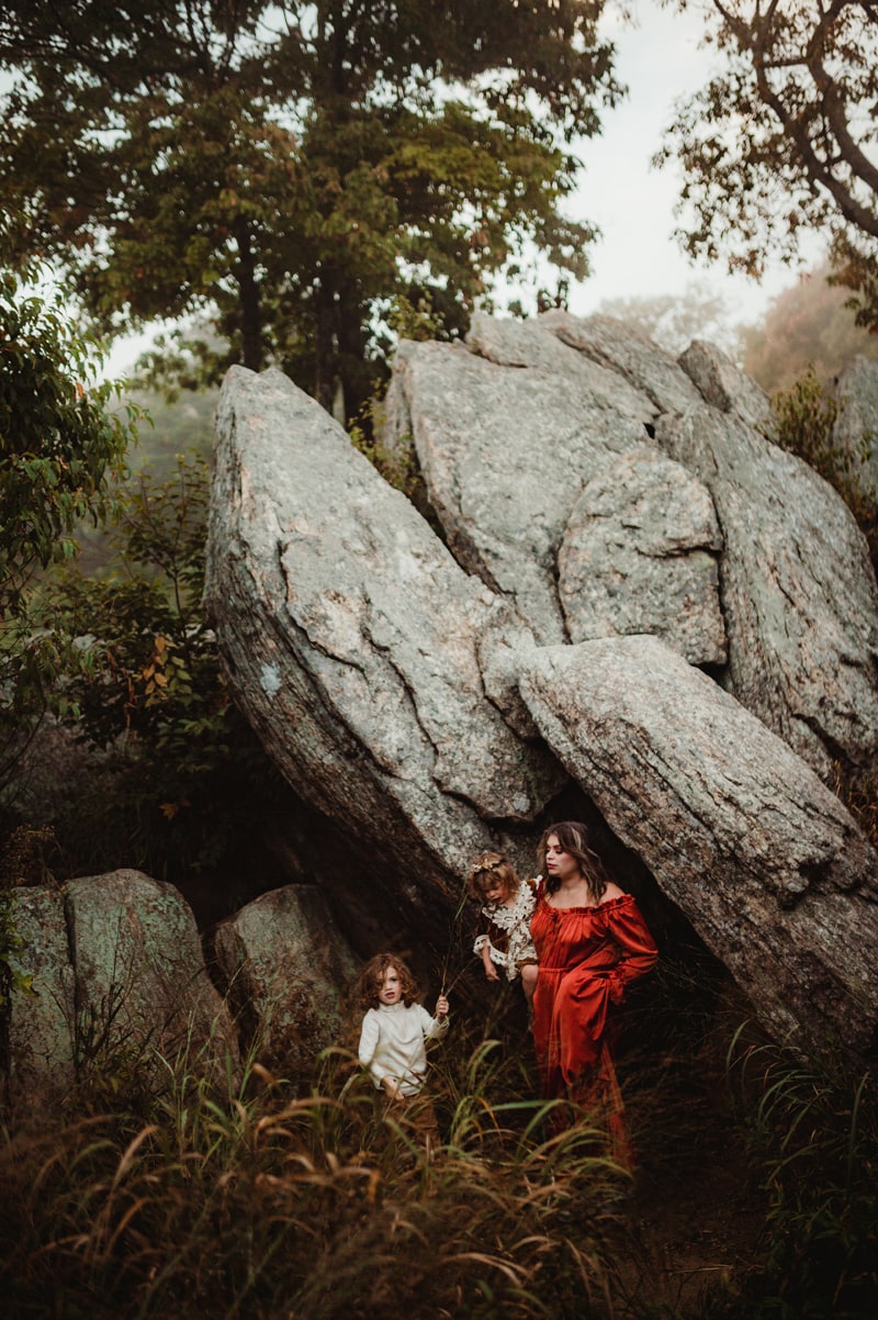 Family Photographer, mom holds baby girl and another daughter stands near them, they are outside near big boulders and lots of vegetation