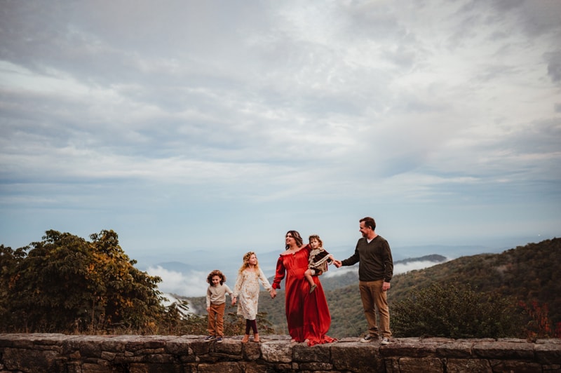 Family Photographer, a family of five stand on an old rock wall before a large forest and a cloud-filled sky
