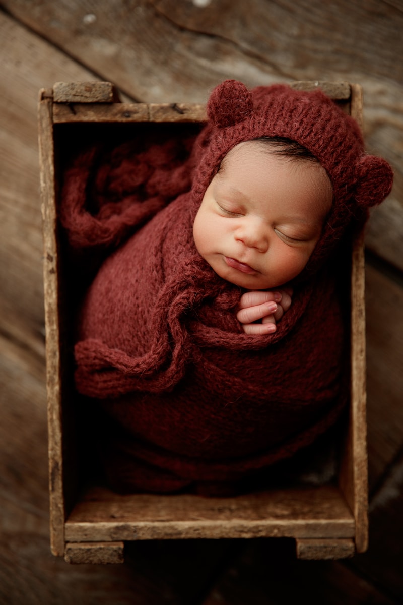 Newborn Photographer, a baby boy is wrapped in knit blankets and reclines in a wooden crate