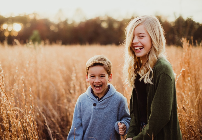 Family Photographer, a sister and brother walk hand-in-hand through a field of tall grass