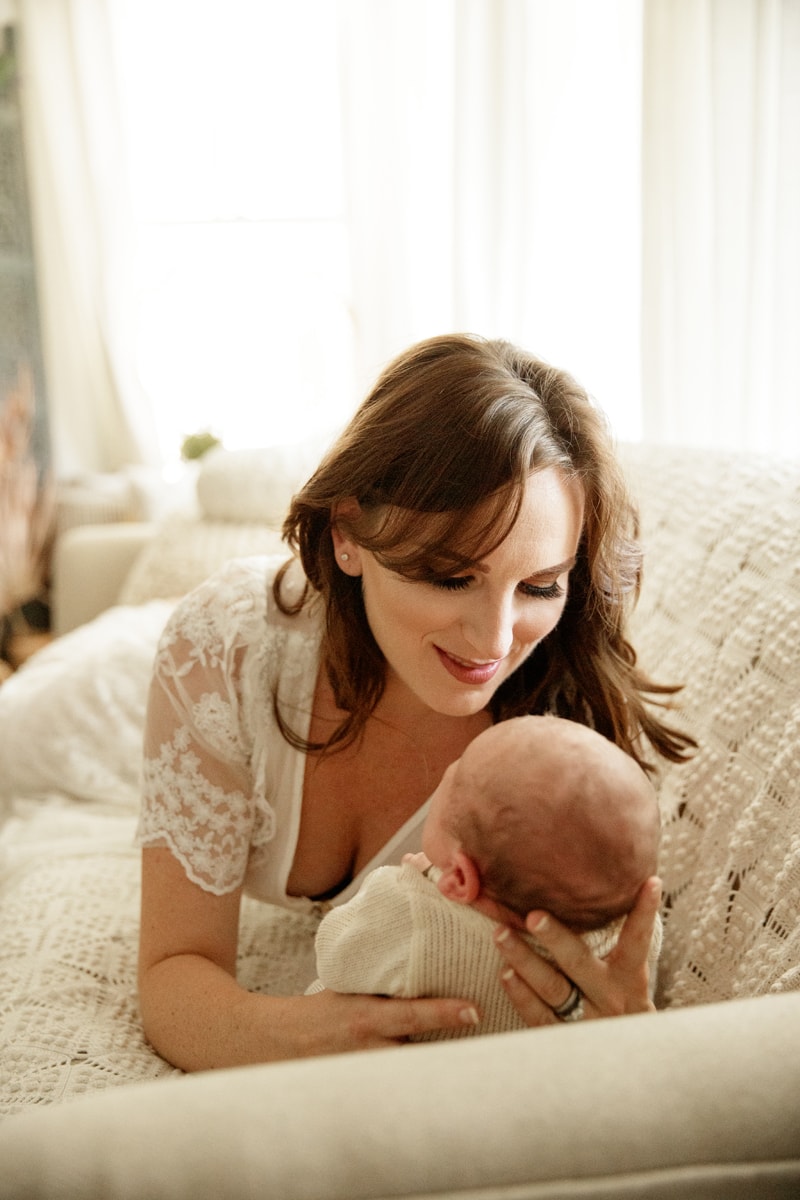 Lifestyle Photographer, mom lays on the couch holding her baby before her. She smiles as she admires him.