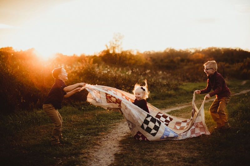 Family Photographer, two older brothers hold a quilt as their younger sisters plays inside of it on a country trail