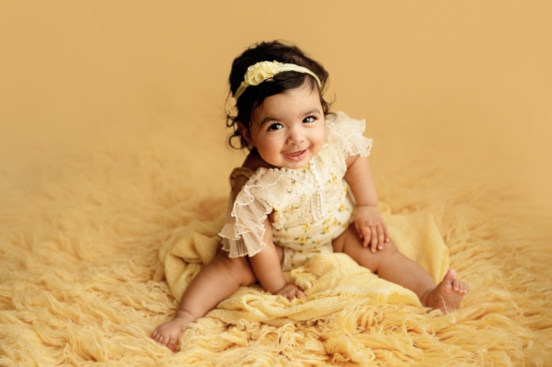 Baby Photographer, a little girl smiles as she sits on plush bedding, head band in her hair