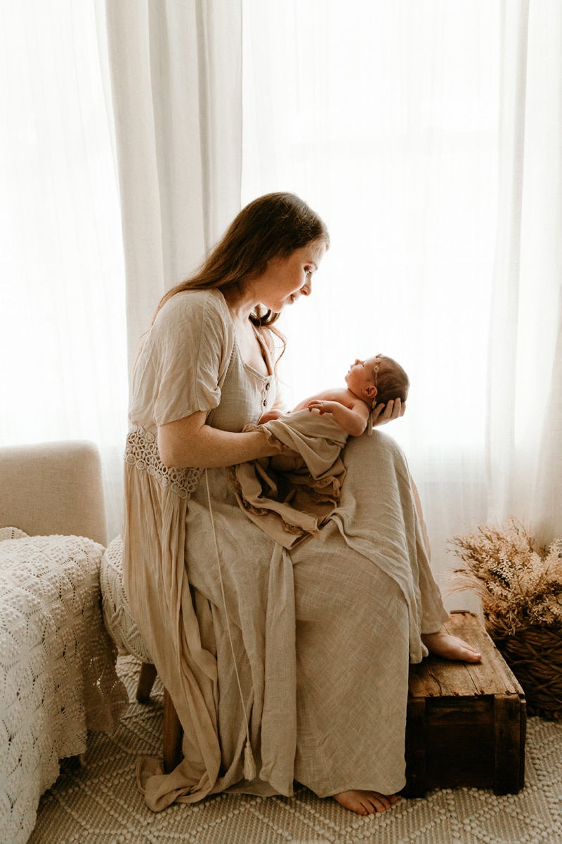 Lifestyle Photographer, a young mother sits beside the bed holding and gazing into baby's eyes