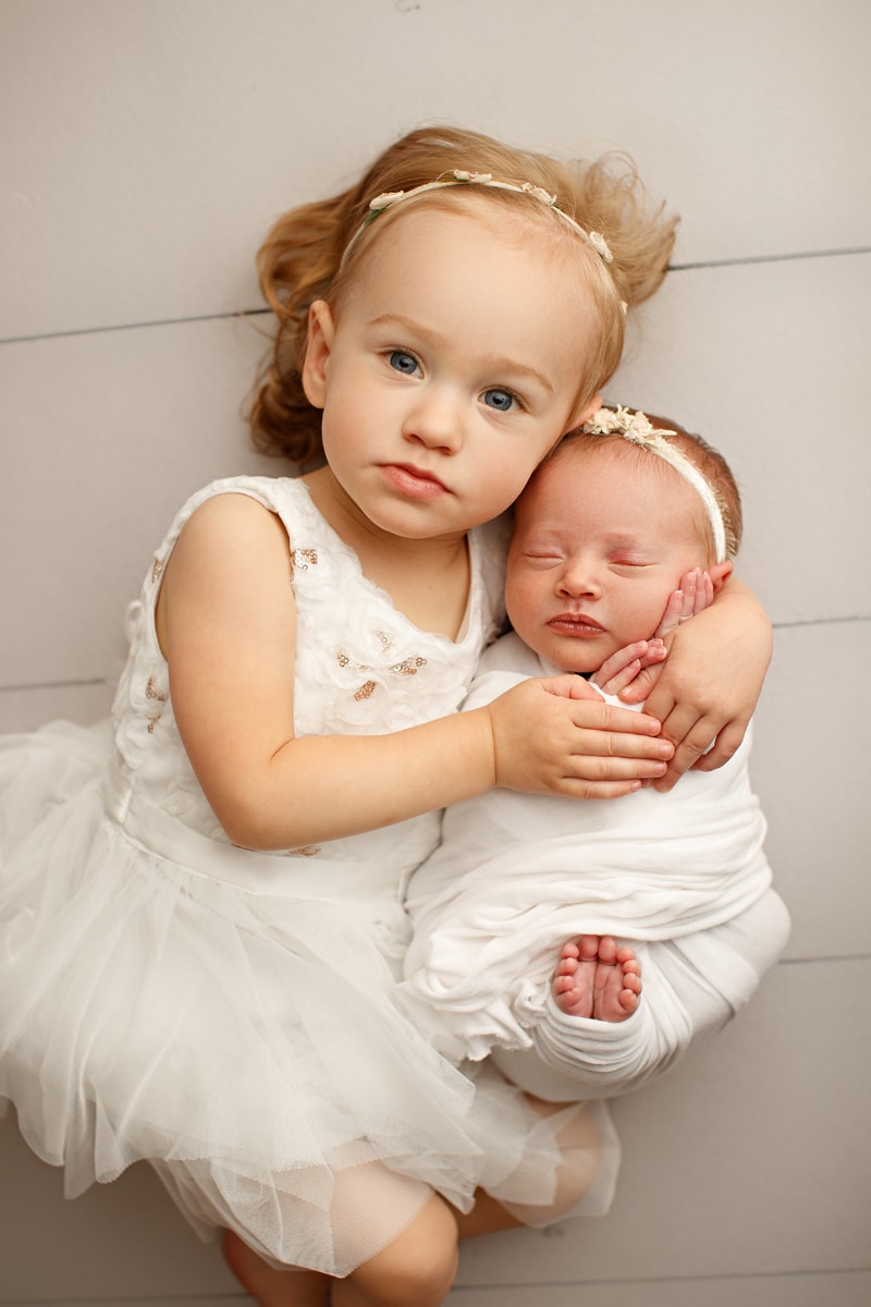 Newborn Photographer, older sister in her dress snuggles newborn baby sister swaddled in blankets