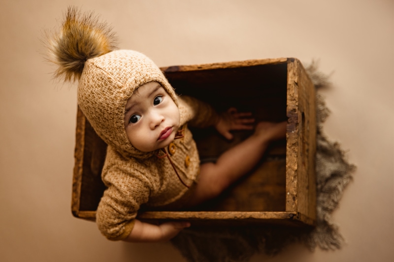 Baby Photographer, a baby child sits in a wooden crate with a onesie that has a raccoon tail on the cap