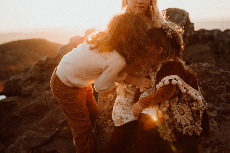 Family Photographer, three young sisters huddle close on a hillside at golden hour