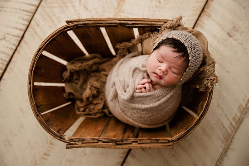 Newborn Photographer, a baby naps cozily in a basket, dressed and bundled in a knit blanket with hood