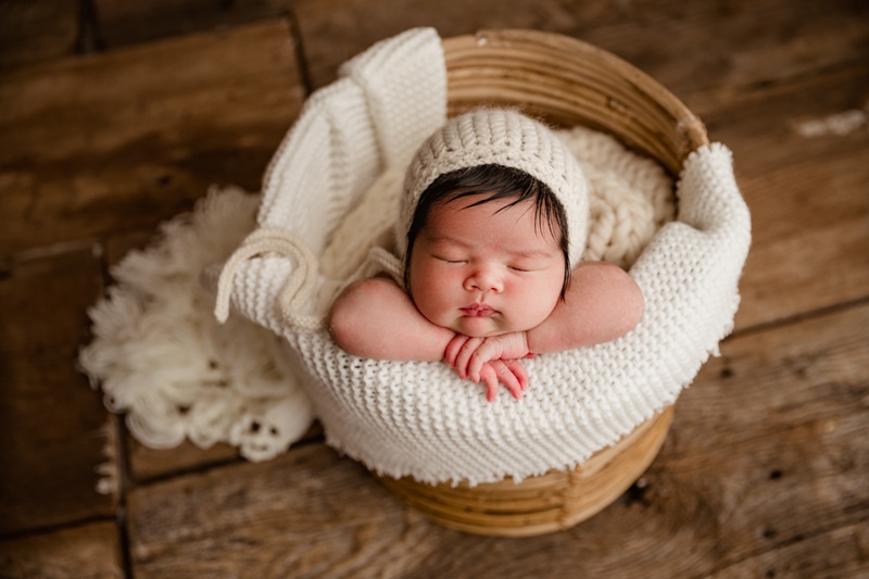 Newborn Photographer, a baby girl lays perched over a baskets edge, wrapped cozy in knit blankets