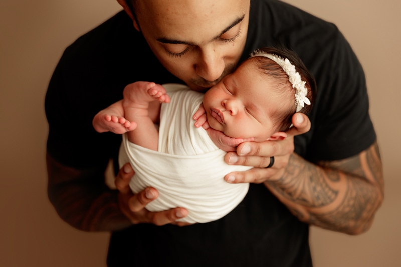 Newborn Photographer, Little baby sleeps as dad holds her and gives her a tender kiss on the cheek