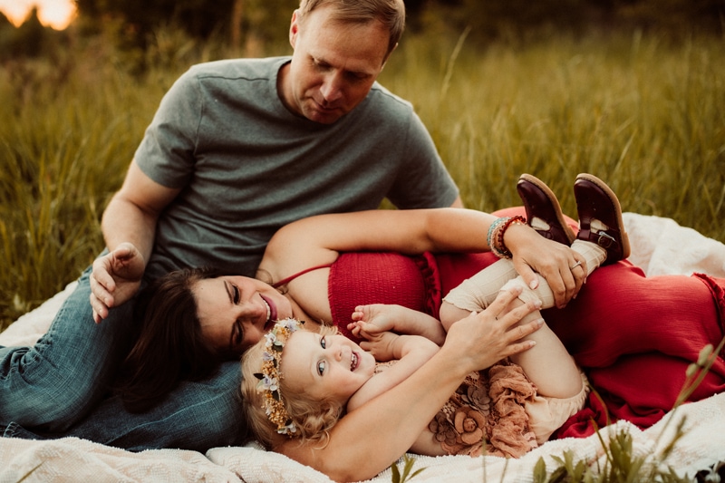 Family Photographer, mom and her baby girl lay on dad's lap on a picnic blanket oustside