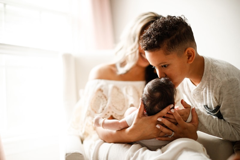 Lifestyle Photographer, mom holds her new baby, older brother comes in to kiss baby on the head