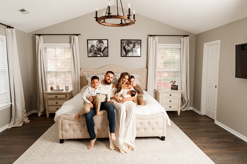 Lifestyle Photographer, Mom, dad, and two brothers sit on a bed celebrating their new sibling in mom's arms