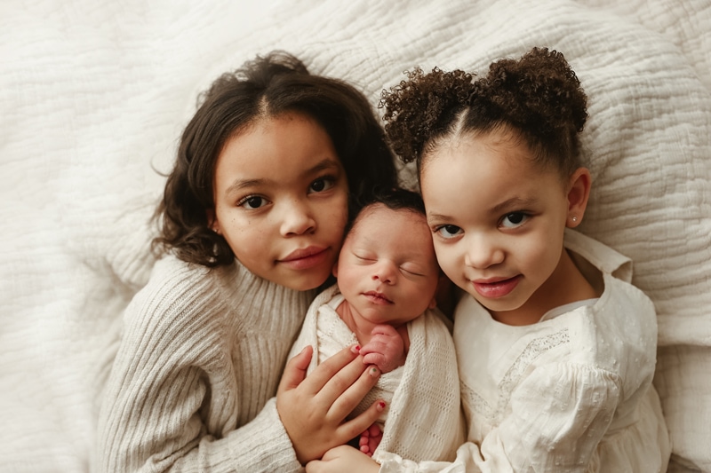 Newborn Photographer, two older sisters hold their new baby sibling close