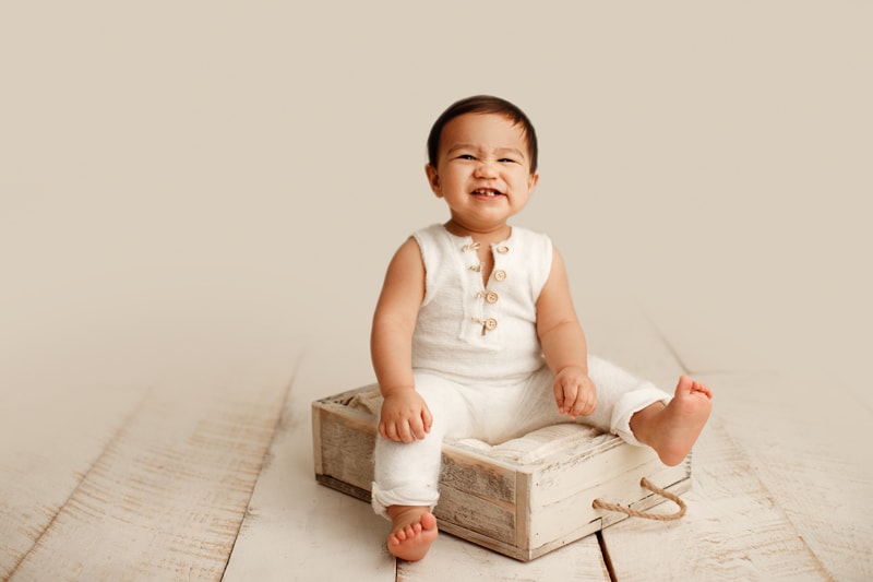 Baby Photographer, a little boy sits on a cushion within a box, he smiles