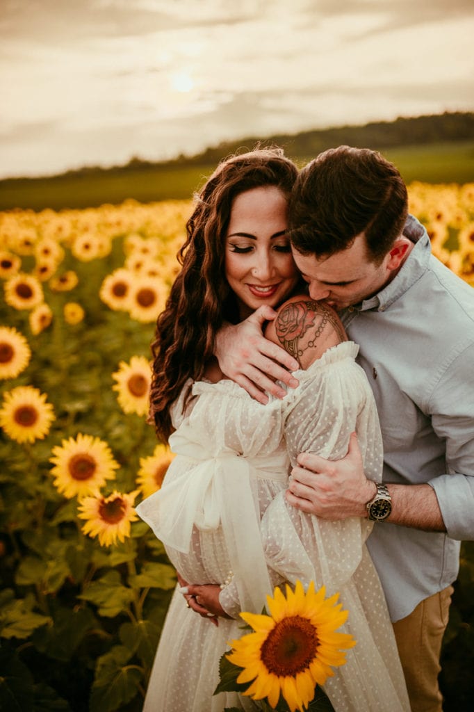 Maternity Photographer, an expecting woman is held by her husband in a field of sunflowers