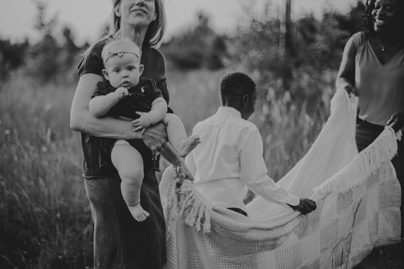 Family Photographer, a mother holds her baby girl as another mother plays with her son and a blanket