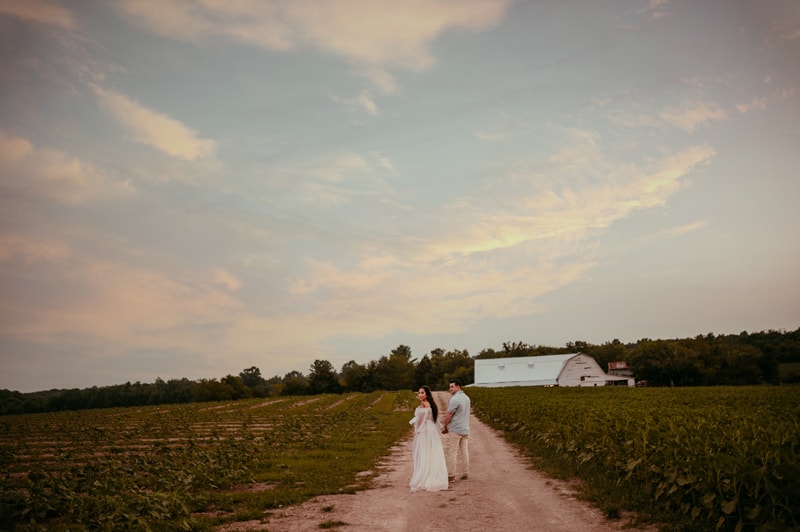Fine Art Maternity Photographer, an expectant mother holds her husband's hands as they walks down a path toward a farm amongst the crops
