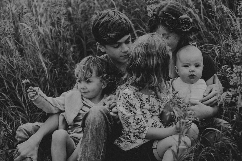 Family Photographer, four older siblings care for baby as they all examine wildflowers as they sit in the grass