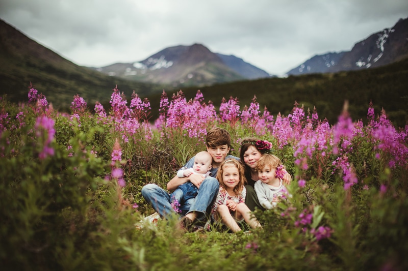 Family Photographer, a teenage boy holds a baby sibling while his three sisters huddle close in a field of flowers before a snowy mountainscape