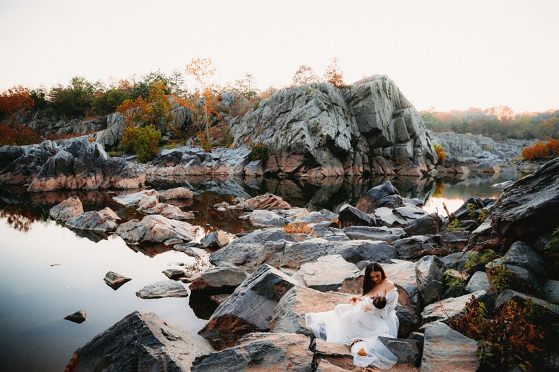 Family Photographer, a woman breastfeeds her baby in a tulle dress near a quiet river filled with large rocks