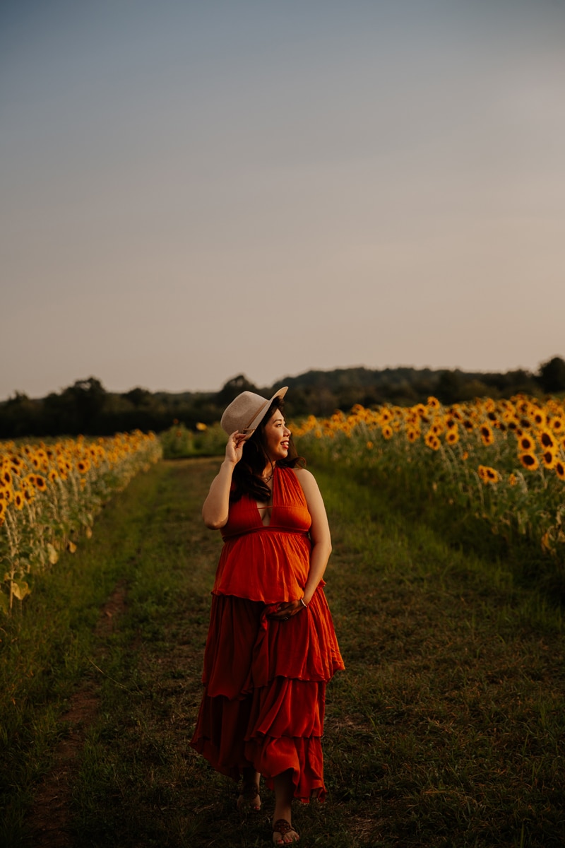 Fine Art Maternity Photographer, an expectant mother stands on a patch of grass between sunflower fields at sunset