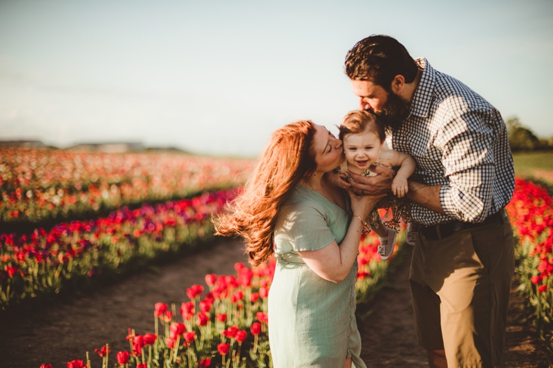Family Photographer, a mother and father hold up and kiss their baby girl in a field of tulips