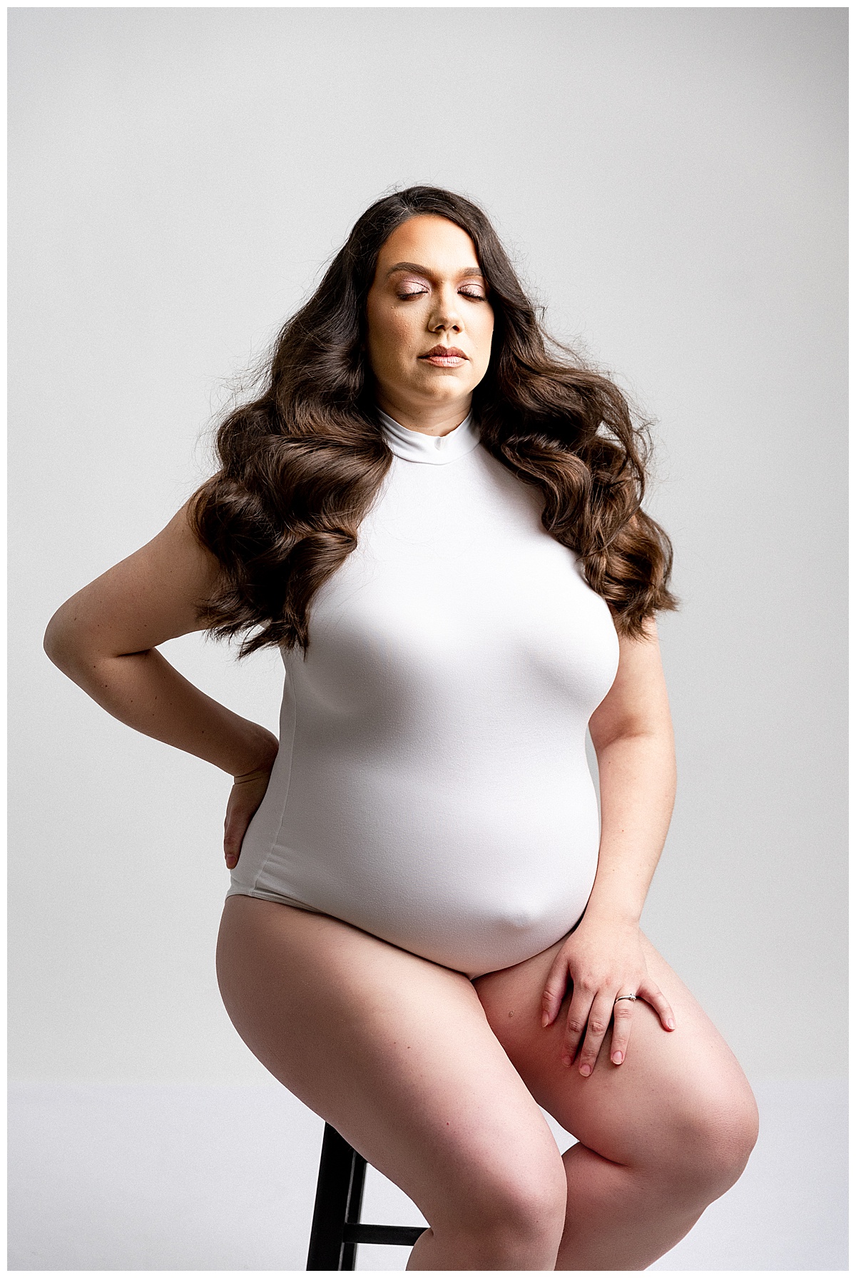 Woman sits on a stool in a white bodysuit for her Full Fine Art Maternity Session